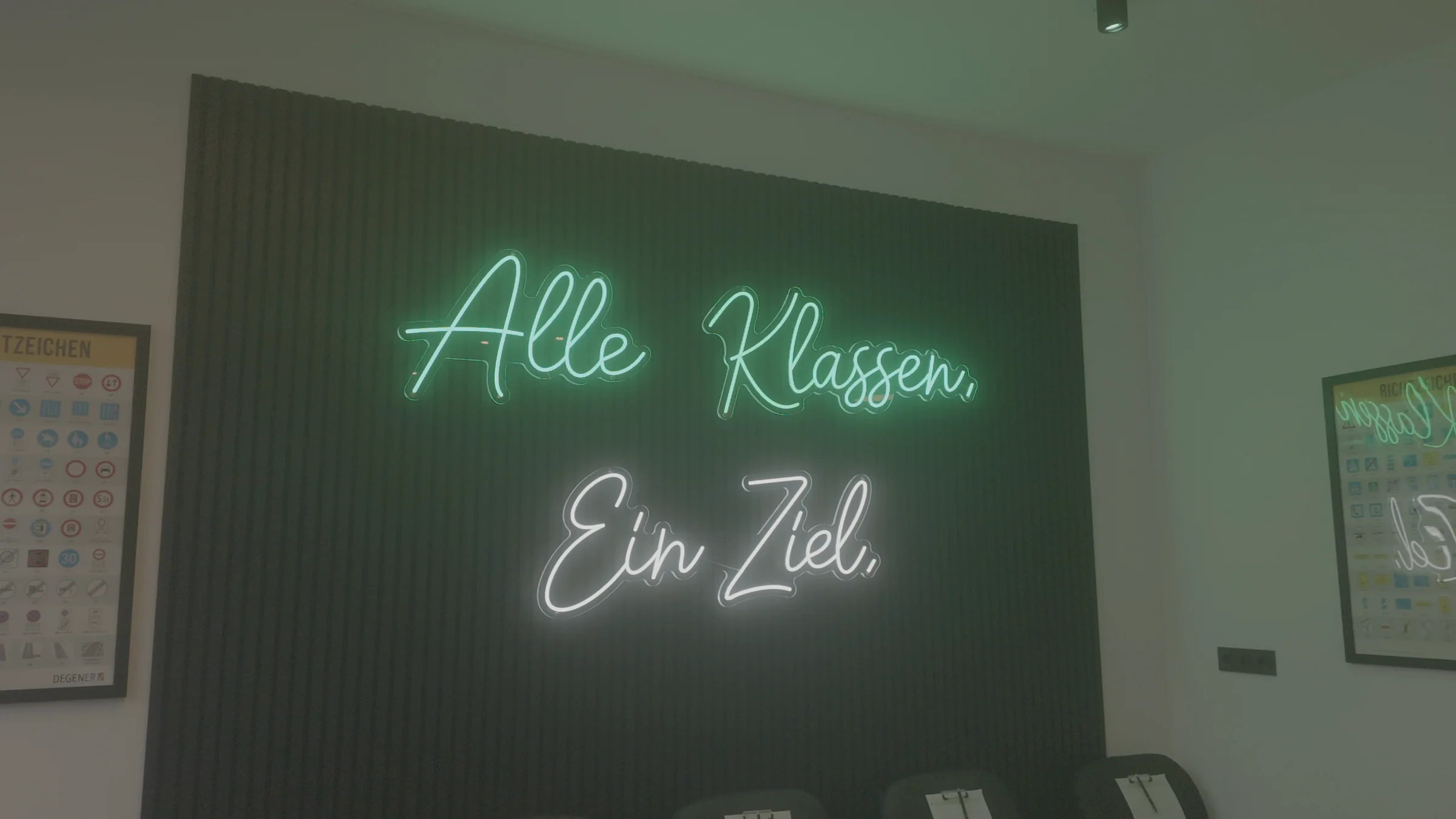 Der Neonschriftzug Alle Klassen, Ein Ziel. leuchtet grün und weiß auf der dunklen Lamellenwand im Theorieraum in Hilden.